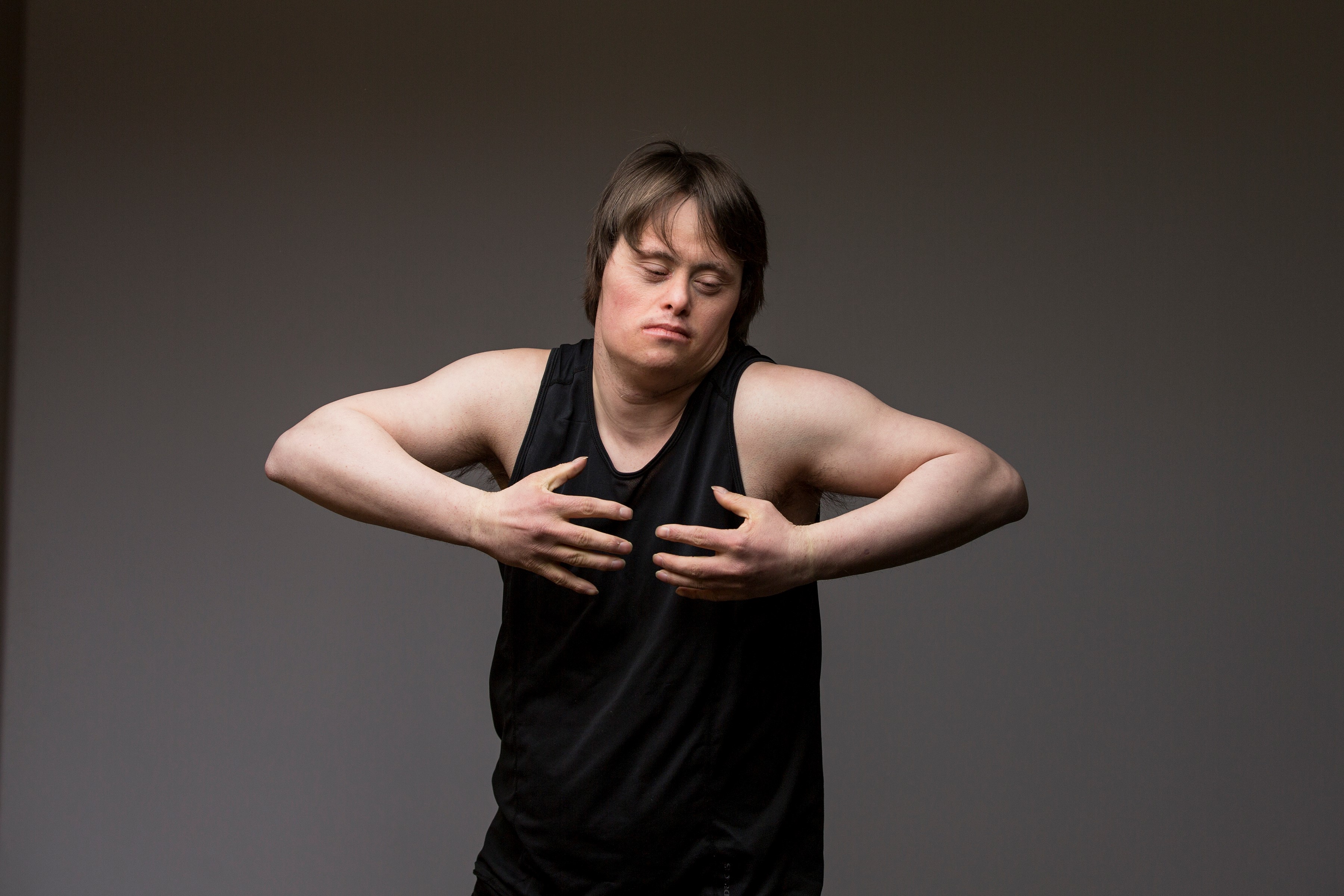 Image description: Luke stands against a grey backdrop. He wears a black singlet, his arms are bent and hands are drawn into his chest. The photo has an emotive physicality to it, like a moment from a dance.