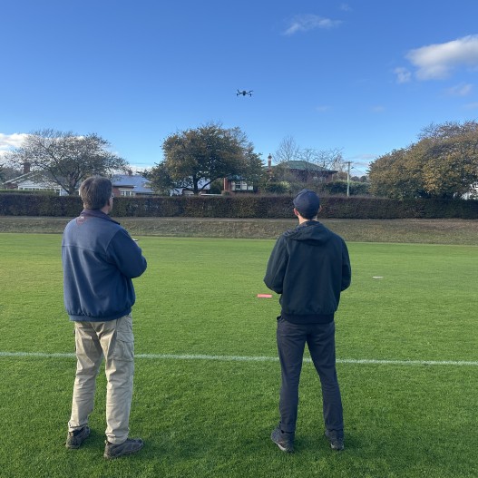 Two people watch a drone flying in a clear sky above a grassy field with trees and houses.