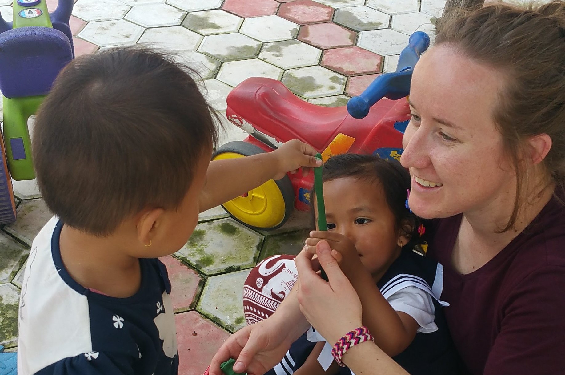 A caucasian woman with light brown hair in a ponytail plays with two Nepalese children, stretching green plasticine between their fingers.