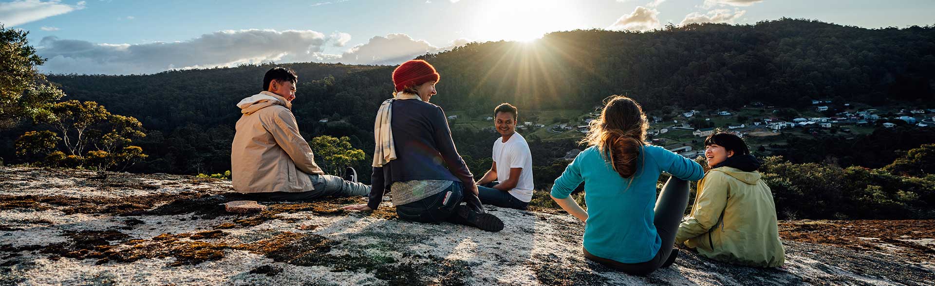 A small group of young adults relaxes on a moss-covered rocky hilltop, gazing at a quaint township below. The sun sets behind a distant hill, casting a warm glow under a clear blue sky.