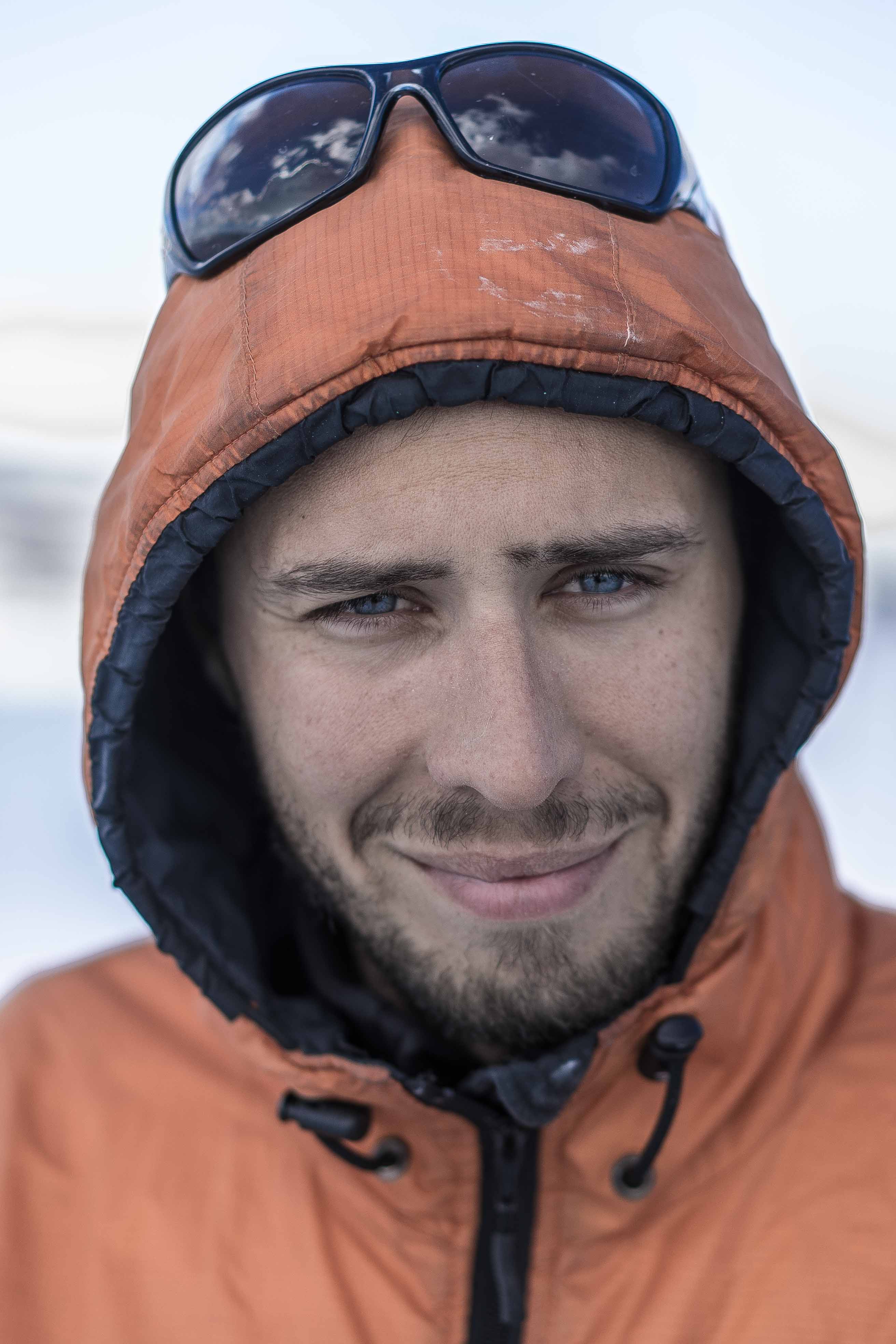Emiliano Cimoli in an orange hooded jacket with black drawstrings and sunglasses on the hood, standing in a snowy or icy environment.