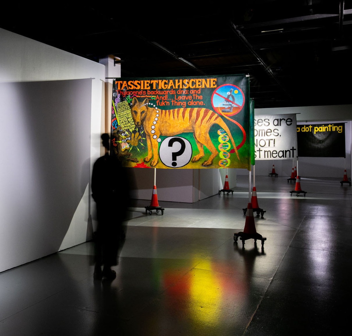 The image shows a dimly lit exhibition space with a shiny, reflective floor. Several large, colorful banners stand freely around the room. The banner in the front features a striking illustration of a Tasmanian tiger, also called a thylacine. The text on this banner says: "TASSIETIGAHSCENE" Thylacine's backwards dna: and ... And ... Leave the fuk'n thing alone." A big question mark is also shown on this banner. In the background, behind this main work, you can see other banners. One of them reads: "Houses are homes, NOT Investment!" and another "a dot painting." The banners are placed on orange traffic cones with wheels, which gives a sense of caution. A person is walking past the main banner, but they look blurry, maybe because they're moving.