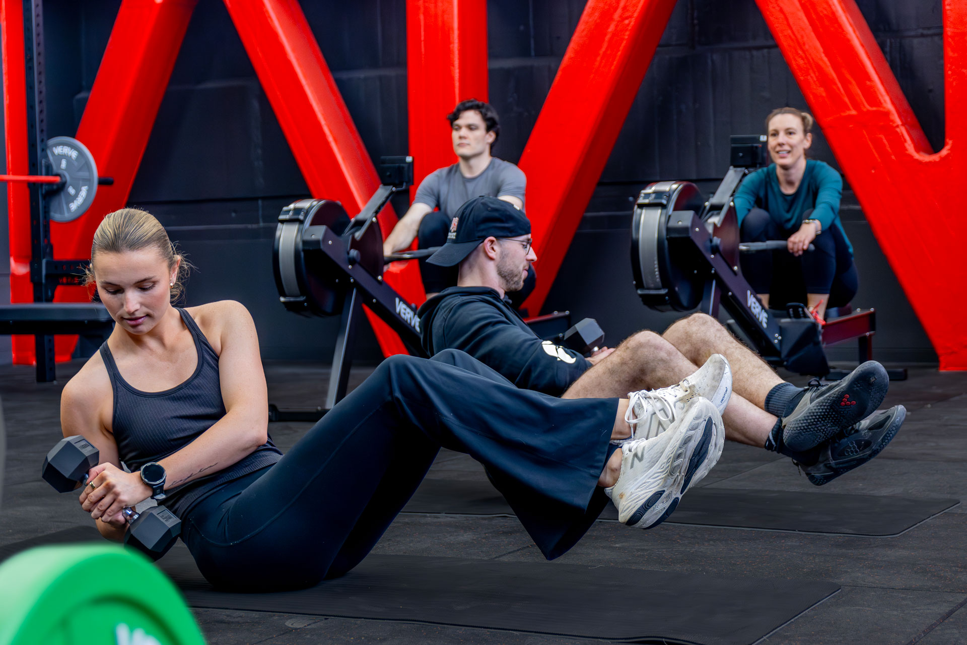 People exercising in gym, one person performing a dumbbell crunch on a mat in the foreground while others use rowing machines behind, set against bold red steel beams.