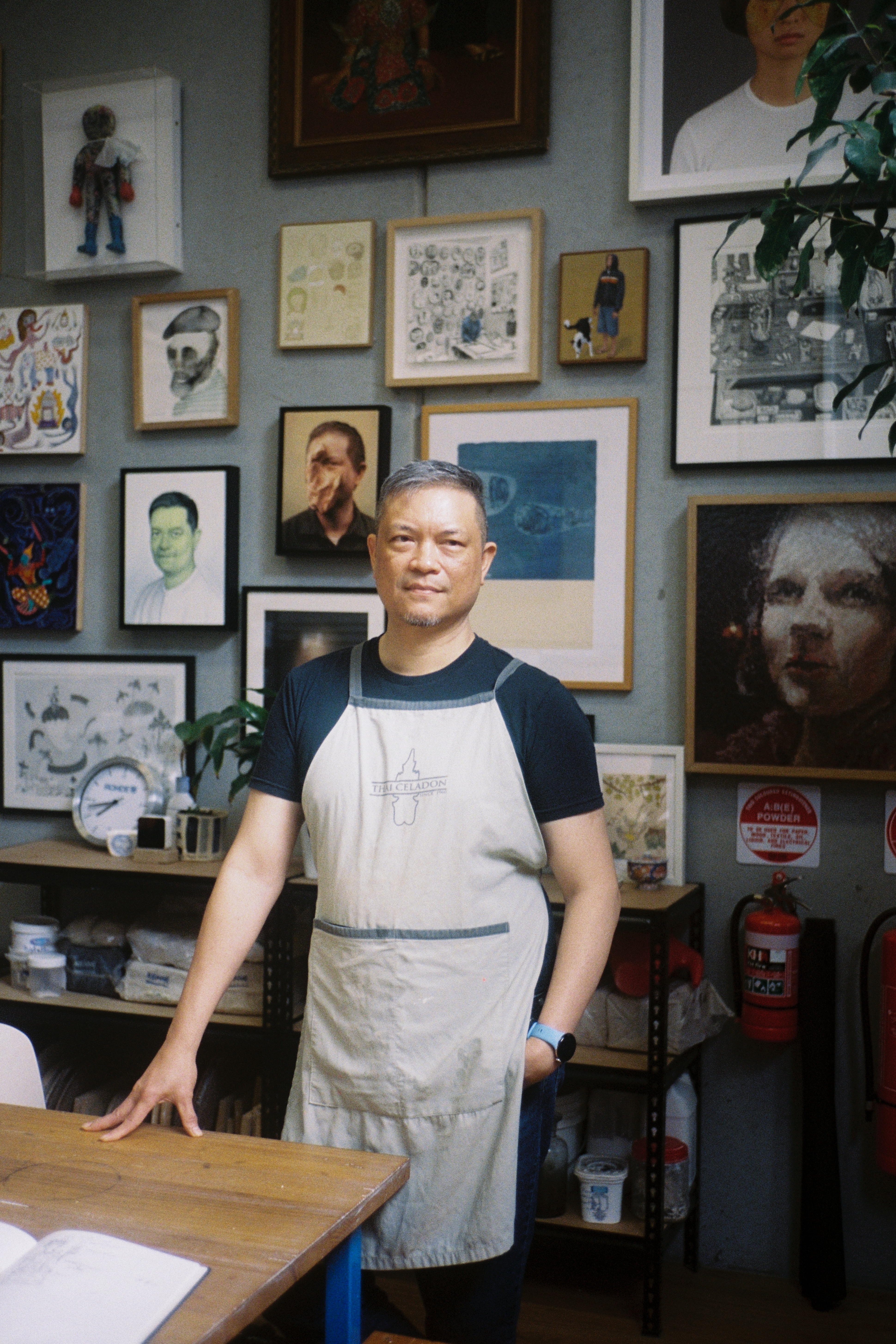 Portrait of Vipoo Srivilasa standing in front of his studio wall filled with framed artworks of various sizes. Vipoo wears a dark and light-coloured T-shirt and faces the camera, while paintings, drawings and photographs create a dense backdrop behind him.