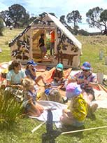 Group of children sitting outdoors in a semi-circle, facing a wooden playhouse, engaged in an educational or recreational activity.