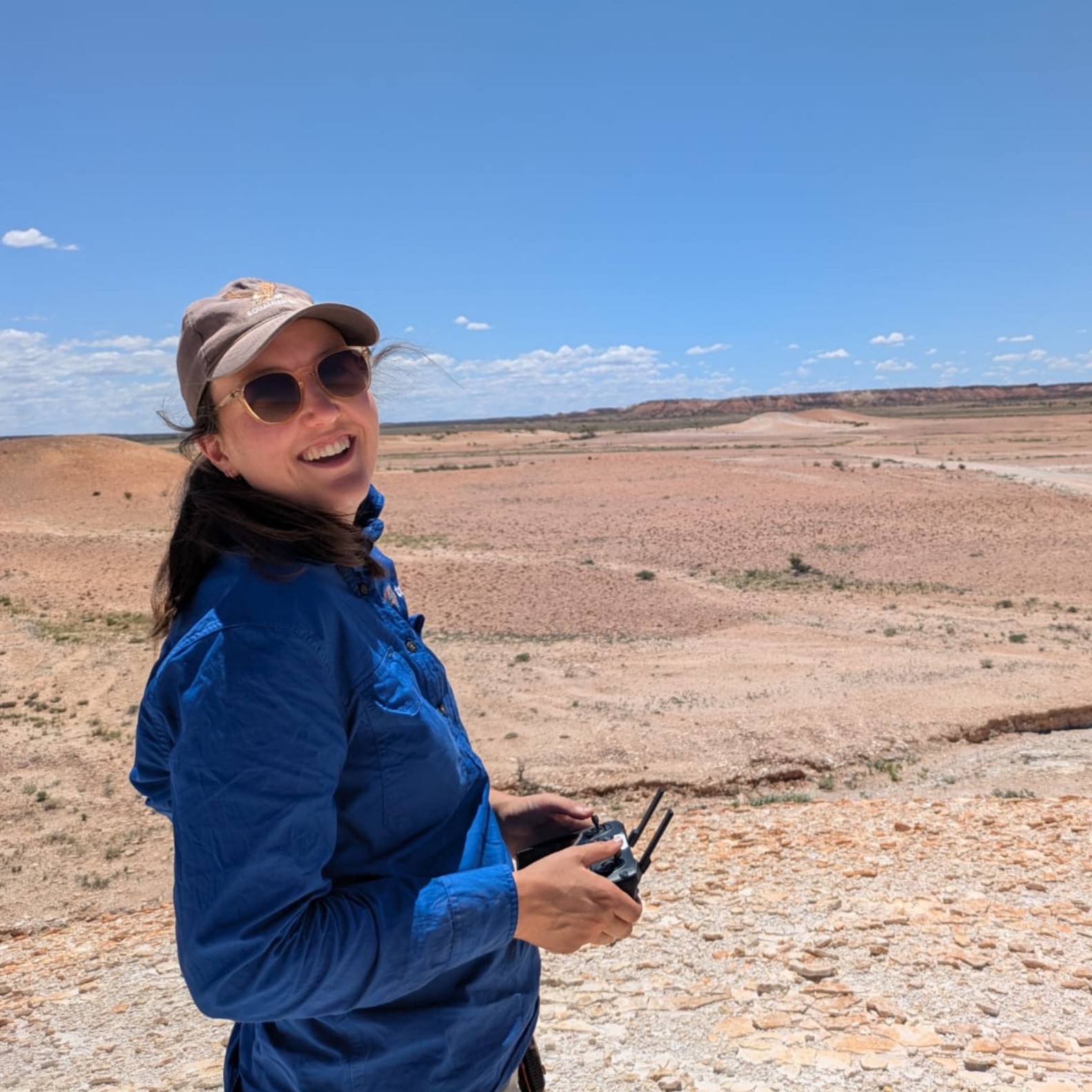 Alice in a blue jacket and cap holding a remote control device in a dry, desert-like landscape under a clear blue sky.