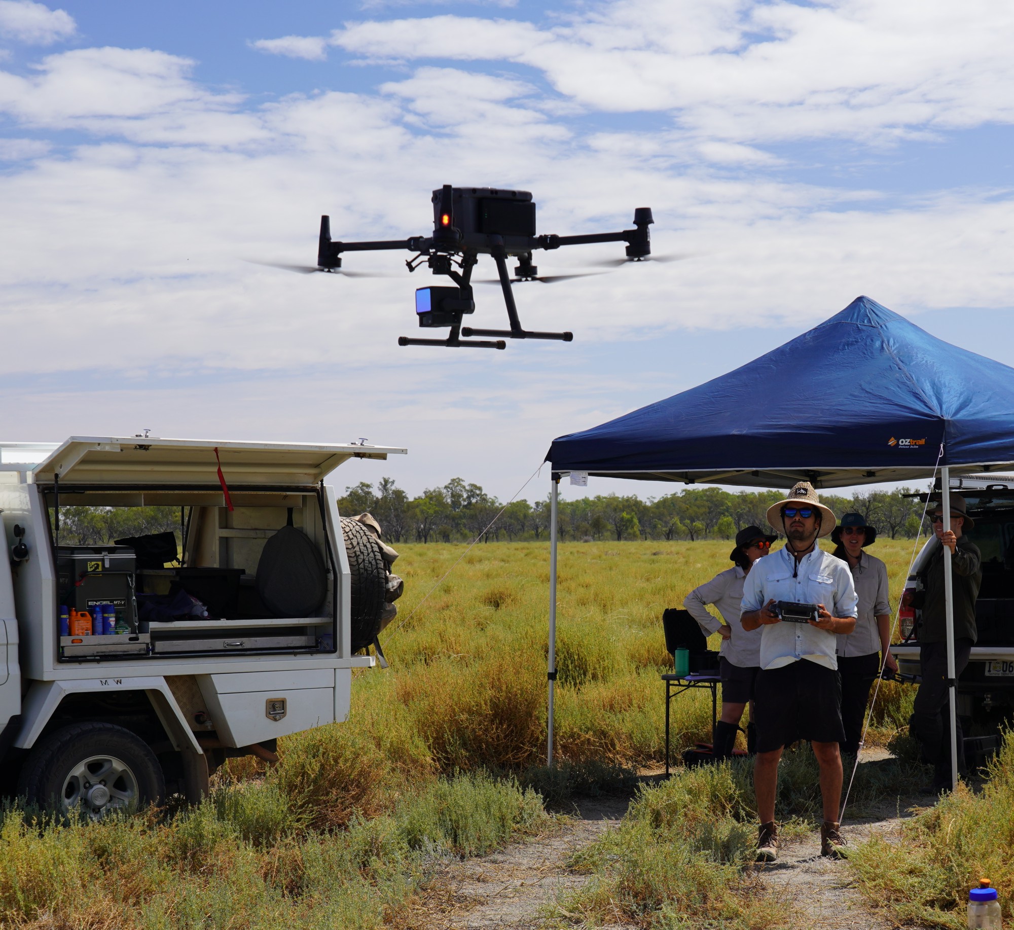 Group operates drone in a field near a white vehicle and blue canopy tent with gear inside.