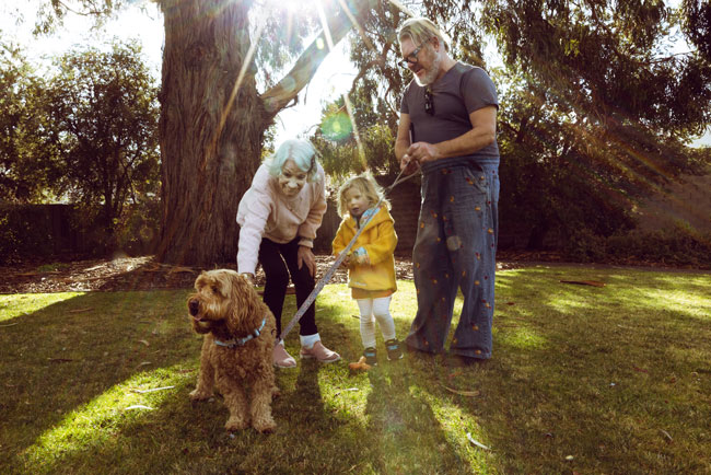Three people and a dog in a sunlit park surrounded by trees and greenery.