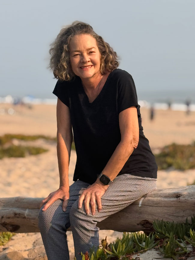 Kim DeBacco sitting on a driftwood log at a sandy beach, wearing a black shirt and patterned trousers.
