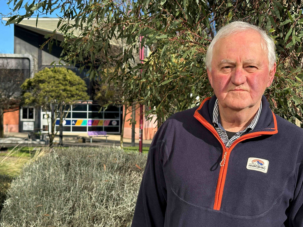 Alistair Scott standing outdoors near campus buildings and trees, wearing a navy fleece with an orange zip.