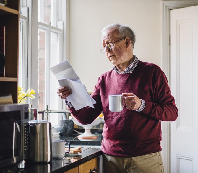elderly man in a kitchen holding coffee cup in one hand and insurance papers in the other