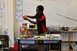 Individual standing in front of a classroom, engaging in an activity on a table with various objects, with educational posters on the wall behind them.