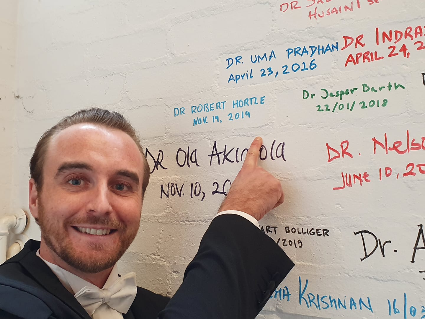 Robert Hortle smiles at the camera, pointing to his name written on a white-painted brick wall, alongside the names of other Oxford students who have successfully defended their theses over the years.