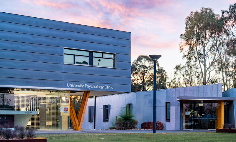Modern University Psychology Clinic with large windows, orange entrance structure, lawn and trees, set against a vibrant sunrise or sunset sky.