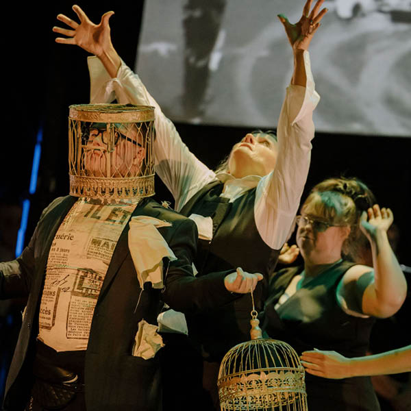 The image depicts a theatrical performance with a surreal and expressive atmosphere. In the foreground, a performer is dressed in a dark suit jacket over a shirt printed with newspaper text, with scraps of fabric protruding from the clothing. A birdcage is placed over the performer’s head like a helmet. Beside this figure, another performer in a white shirt and dark vest stands with arms dramatically raised toward the sky, suggesting a gesture of release or exaltation. This second performer holds an ornate birdcage by its handle. In the background, additional performers are partially visible, and a large grayscale projection adds depth to the scene. Warm stage lighting highlights the costumes and props against a dark backdrop.