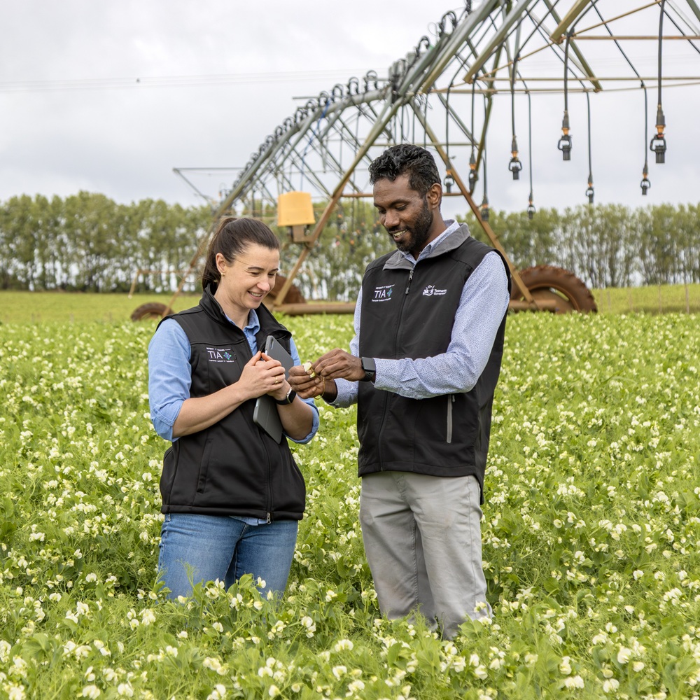 Two people stand in a paddock with an irrigator in the background.