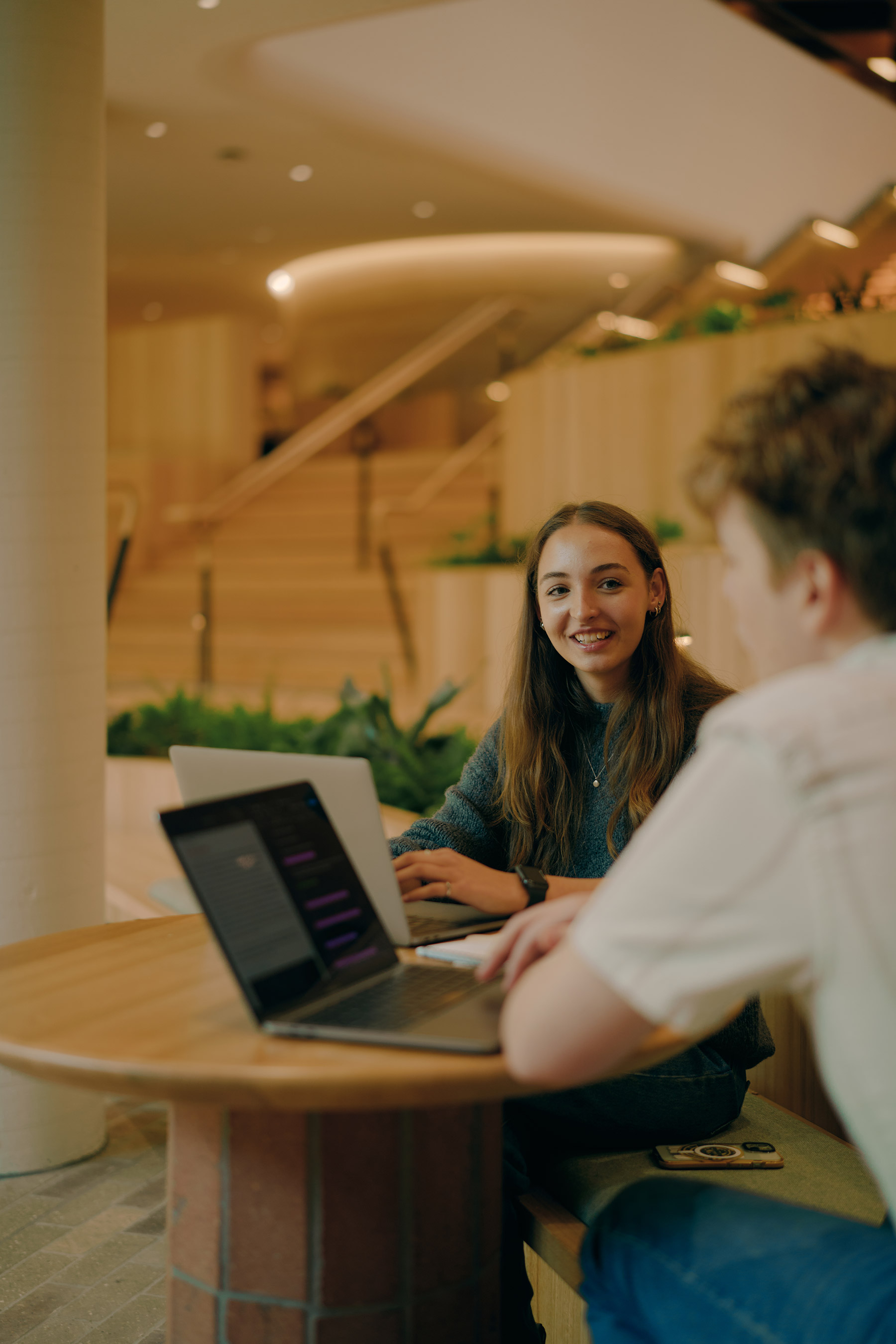Students seated in the new Forestry building using a laptop, with a staircase and warm ambient lighting in the background.