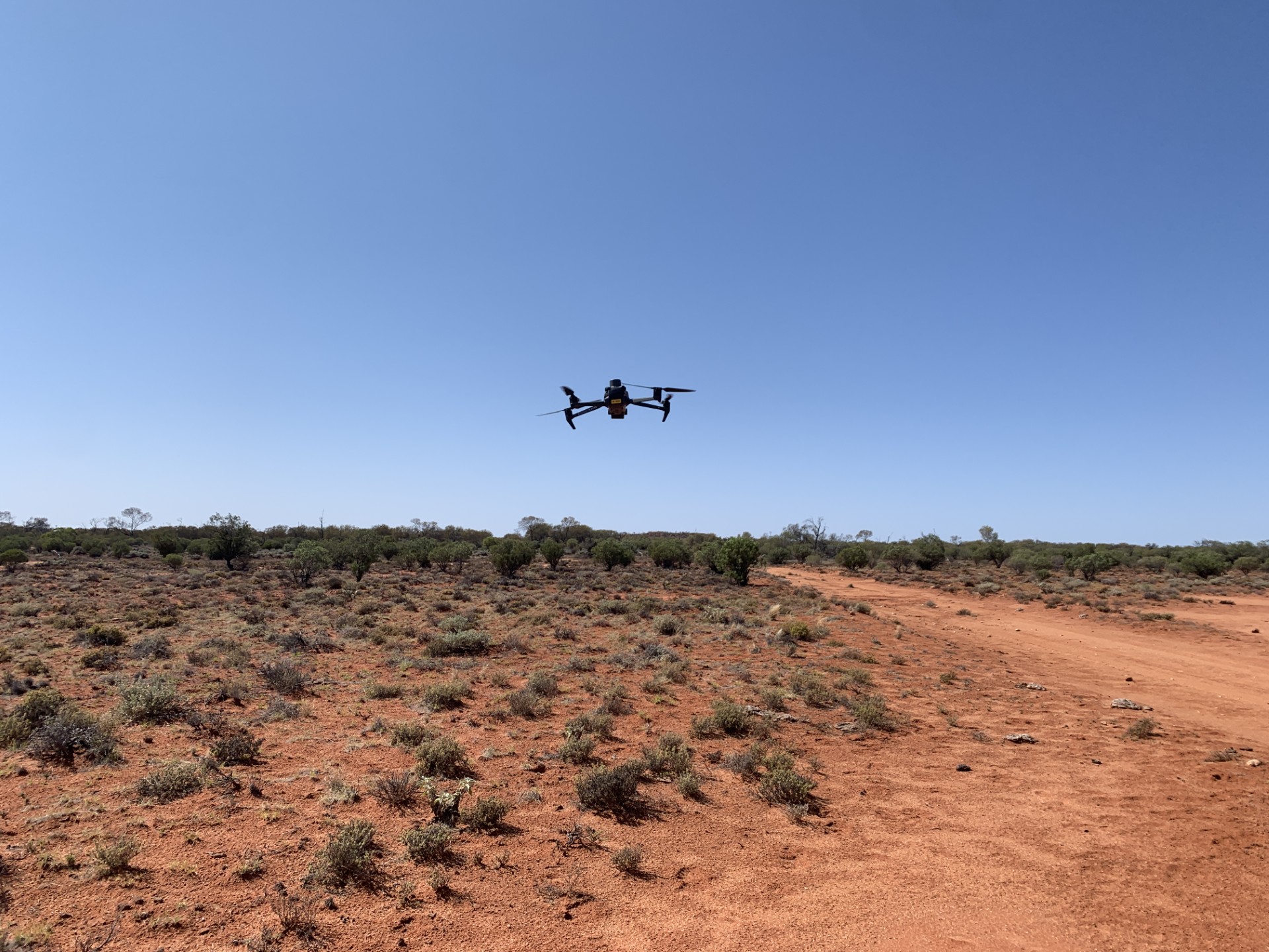 Drone flying over dry, arid terrain with sparse vegetation and a dirt path under a clear sky.