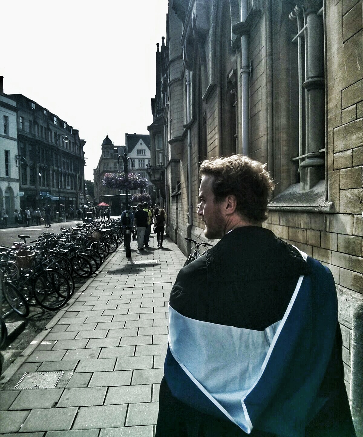 Robert Hortle, his back to the camera, is wearing a black academic gown with a pale blue hood hanging below his shoulders as he walks along a street lined with historic buildings in Oxford, UK.