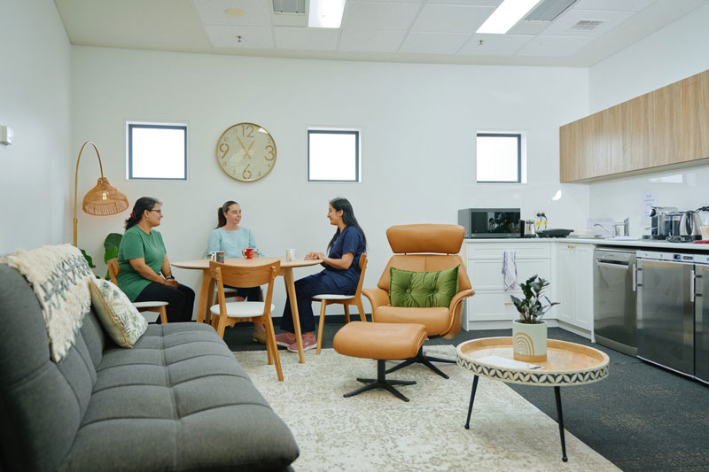 Staff lounge with a grey sofa, coffee table, and armchair in the foreground. Three people sit at a wooden table near a wall clock, with a kitchenette featuring white cabinets and appliances in the background.
