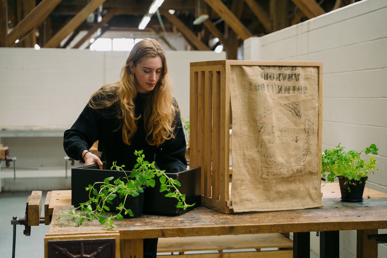 A young woman dressed in all black, with fair skin and long dark-blonde hair, holds a planter overflowing with green herbs as she assembles a sharing-point for fresh produce, which resembles a wooden crate with a hessian sheet over the front.