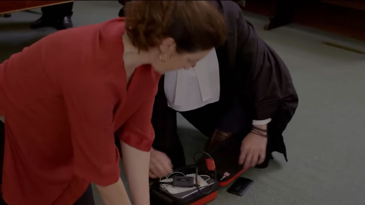A person is performing CPR while another sets up a defibrillator on the floor.