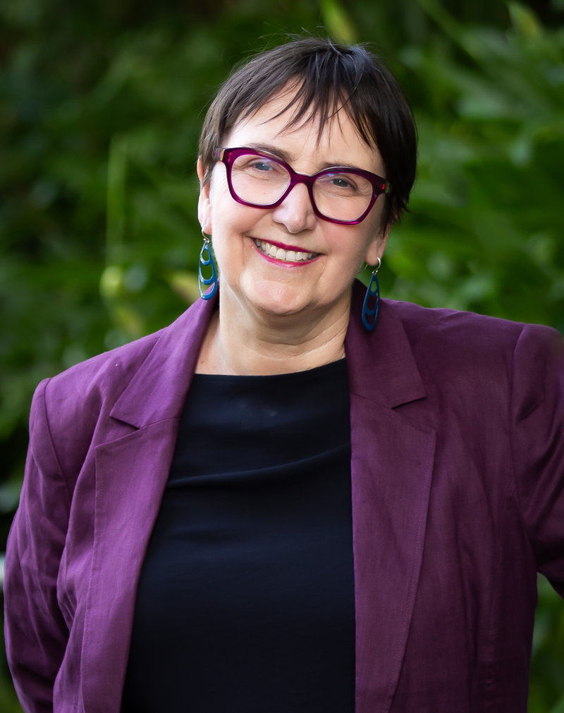 Helen Burnet wearing a purple blazer and dark top standing outdoors in front of green foliage.