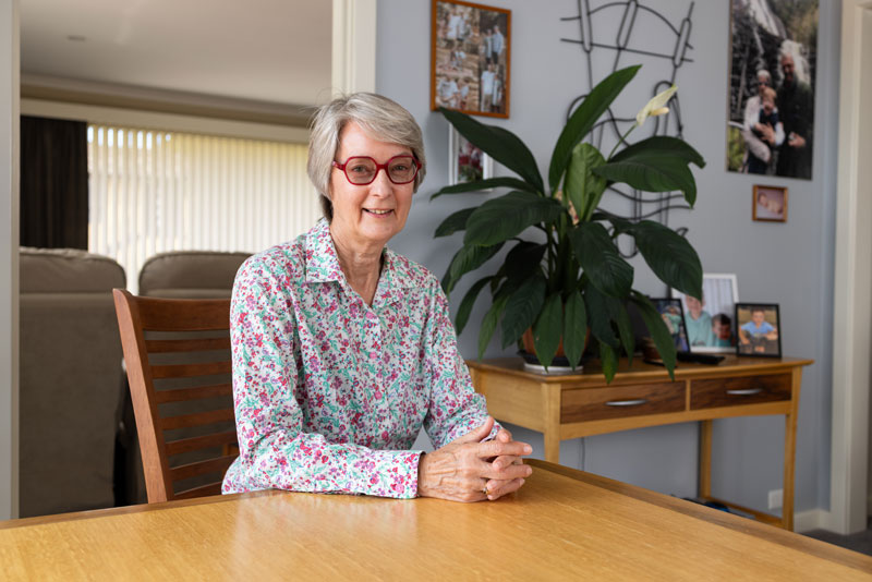 Person seated at a wooden table in a home setting, with indoor plants and framed family photographs in the background.