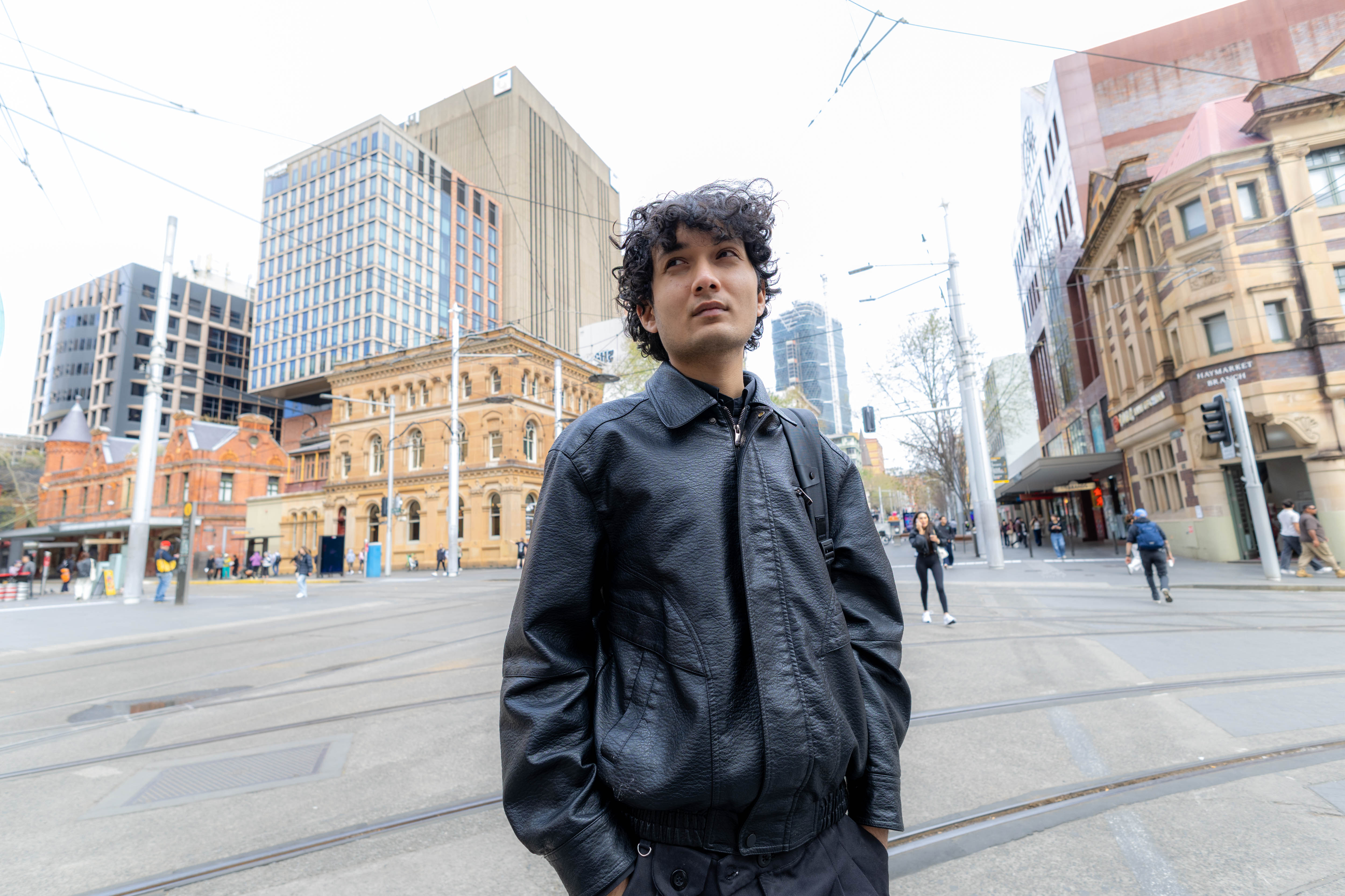 A wide-angle photo of a young Nepalese man, with brown eyes and black curly hair, standing in a Sydney street. He is looking beyond the camera with a slight smile. He is wearing a black leather jacket, the strap of a black backpack over one shoulder. In the background is a large intersection with pedestrians crossing the road, surrounded by city buildings.
