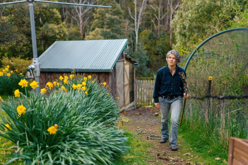 Woman in a blue green flanelette shirt and blue jeans with a rake walking in her garden