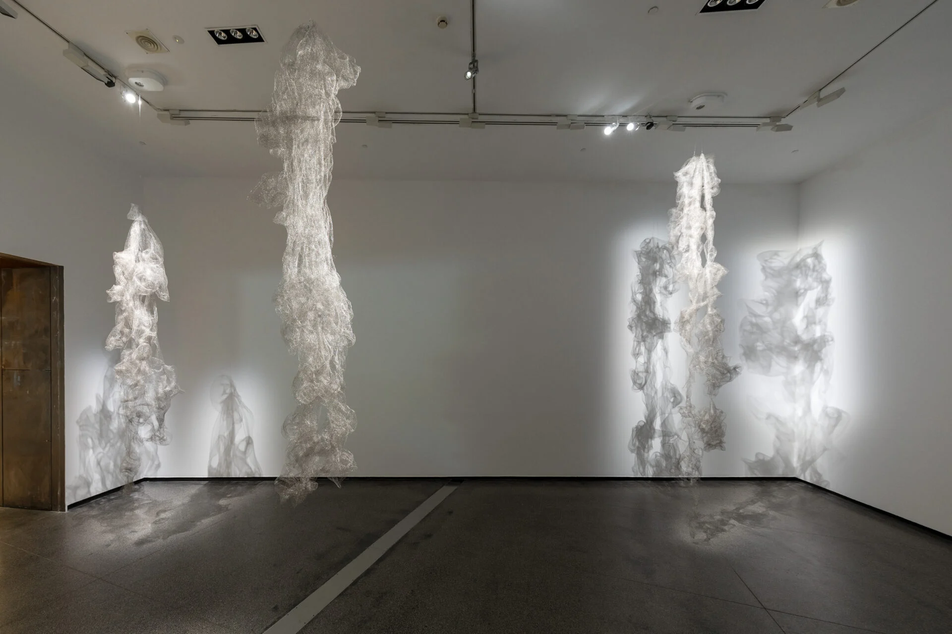 Image Description: Three wire mesh forms hang from the ceiling. The gallery setting features white walls, a lighting track on the ceiling and a grey tiled floor. The cool lighting casts shadows of the mesh forms onto the walls and floor.