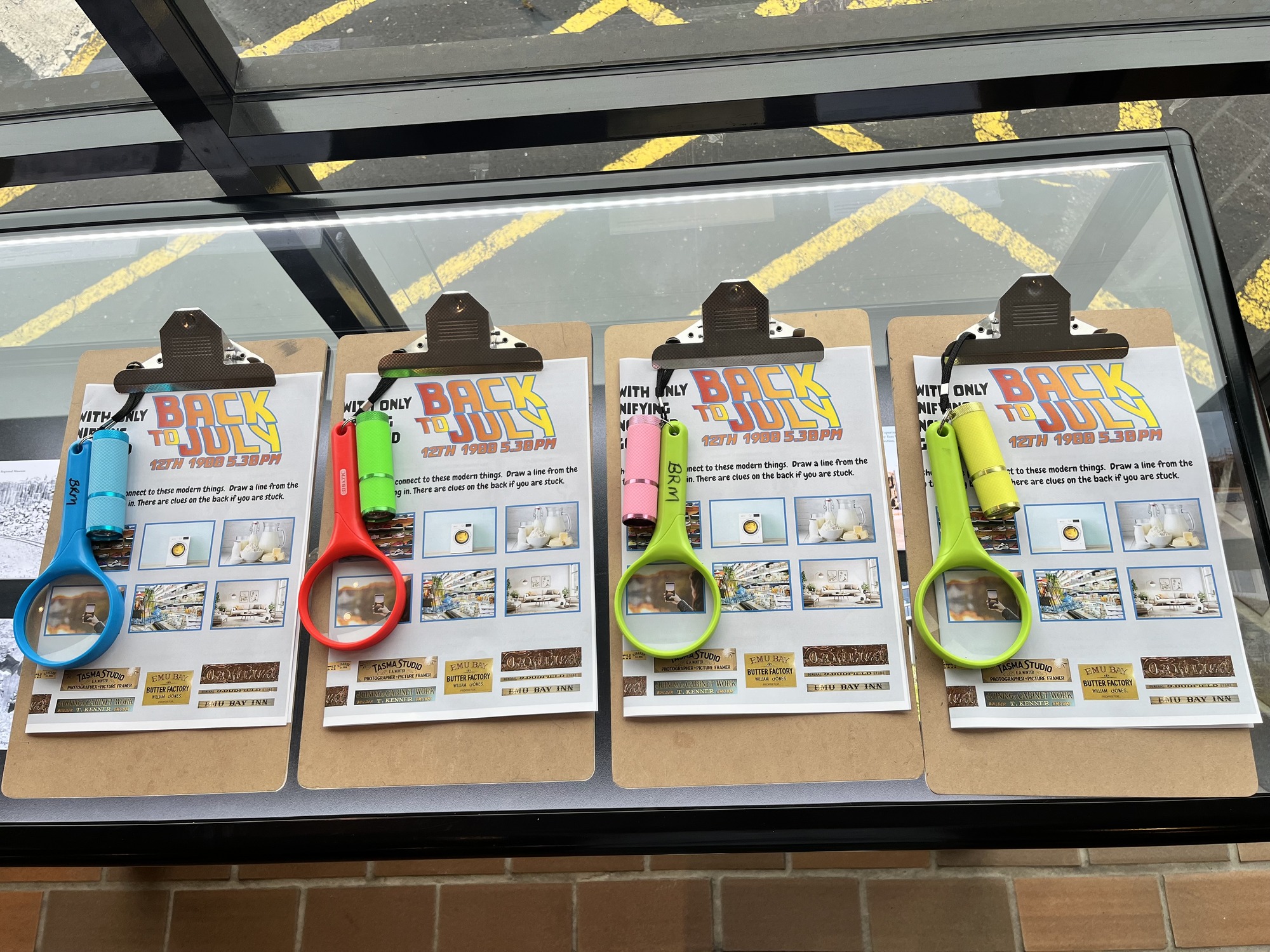 Four clipboards sit on top of a table, each with an activity print-out called 'Back to July' clipped in to place and a magnifying glass and torch hanging from the board. The activity print-out is folded over so not all of it can be seen, but it indicates that the person taking part in the activity needs to follow clues and find things.