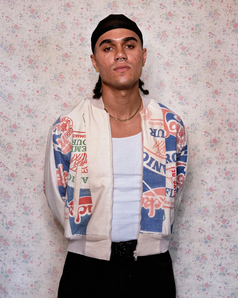 The image features an individual standing against a backdrop with a floral pattern. The individual is wearing a zip-up jacket adorned with various patches and logos. These patches seem to represent different countries.. The colours on the jacket are vivid, including reds, whites, blues, and other hues, making it stand out against the more muted background