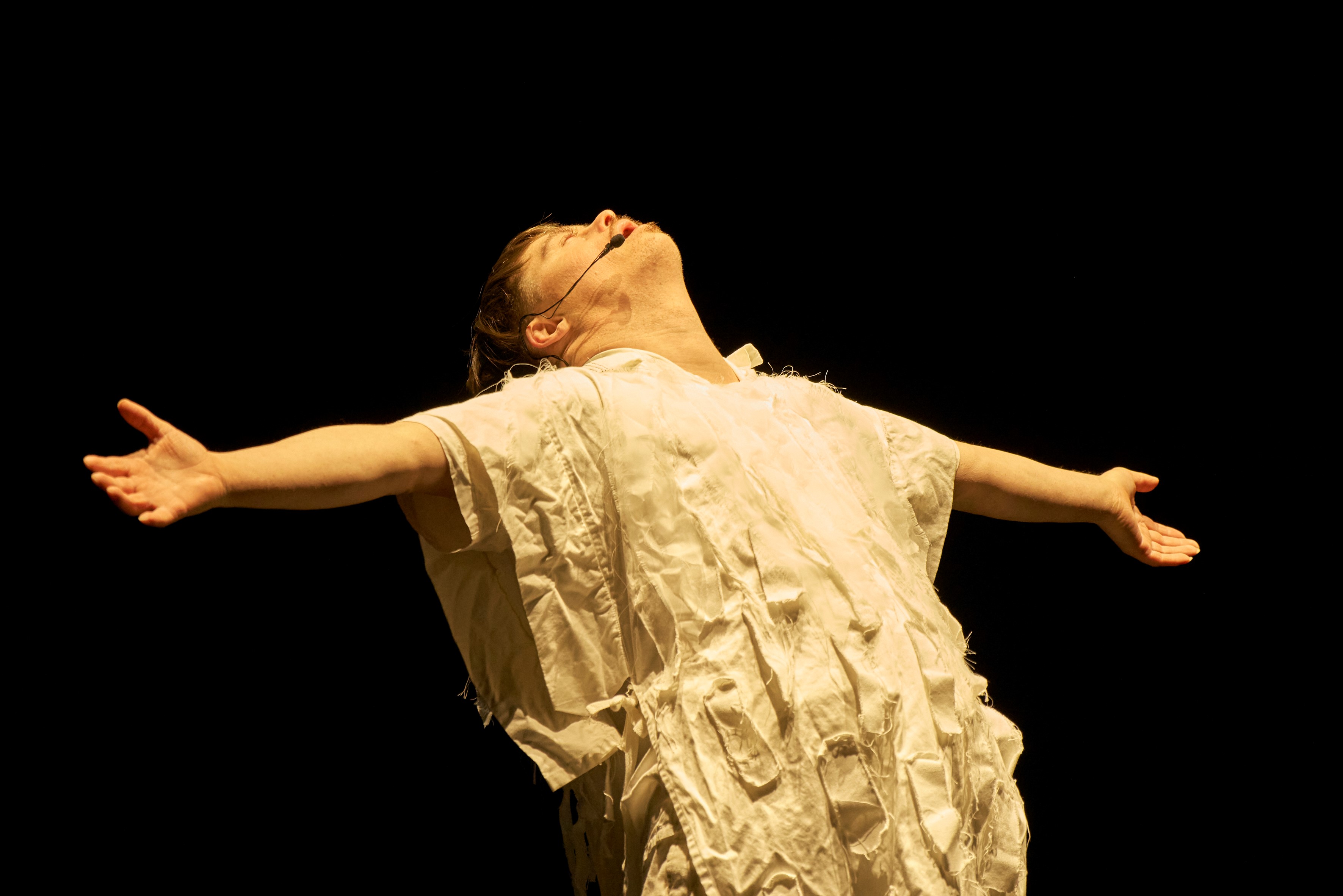 The BOND The image portrays a person (Luke John Campbell) standing on a stage, seemingly engaged in a dramatic performance. Their head tilts back, eyes closed, and arms stretch wide, suggesting an expression of release or embracing the moment. The individual wears a textured, layered garment resembling a dress or costume with ruffled elements. The background is entirely black, emphasizing the figure as if illuminated by a spotlight.