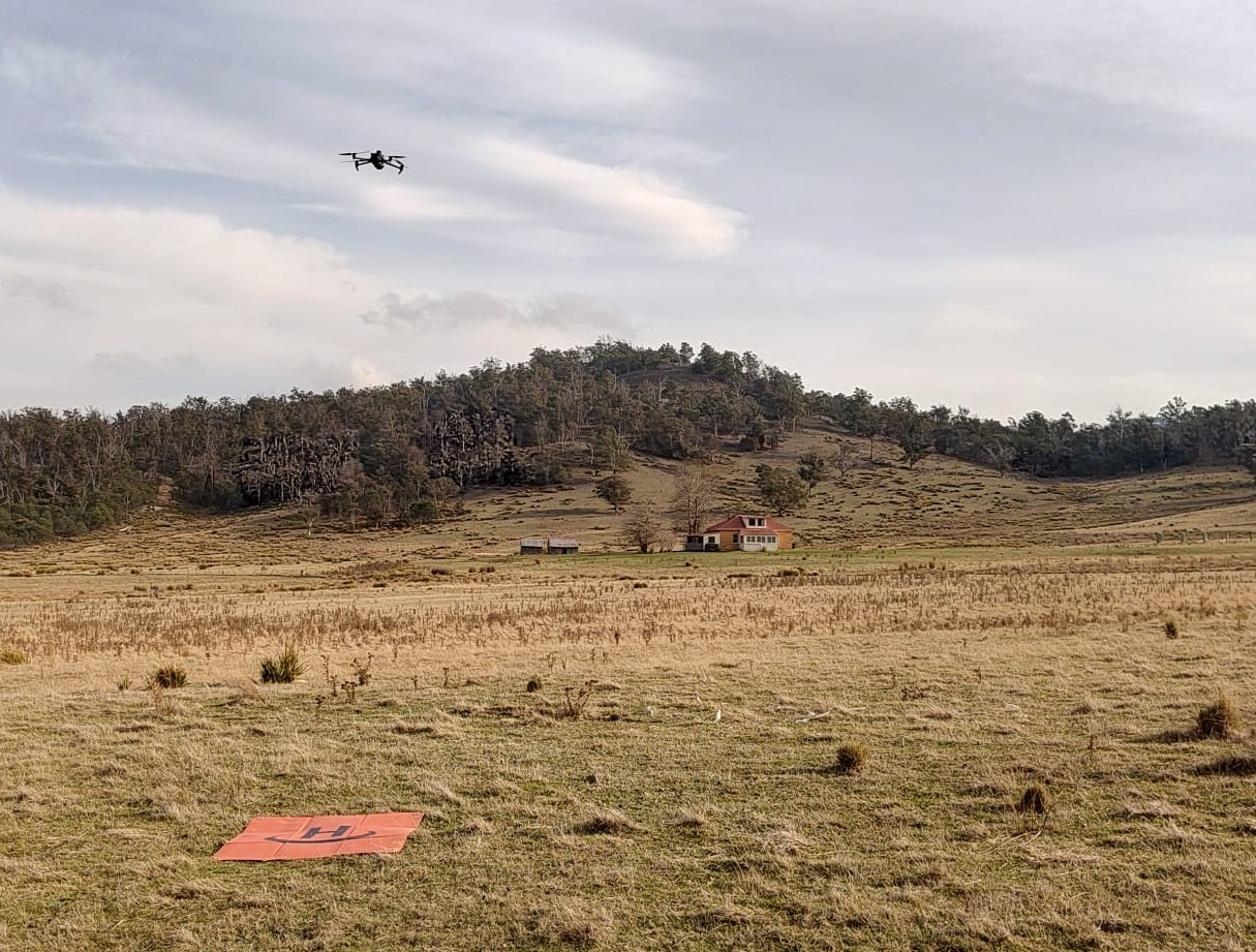 Drone flying over grassy field with trees, a house, and an orange landing pad marked 'H'.