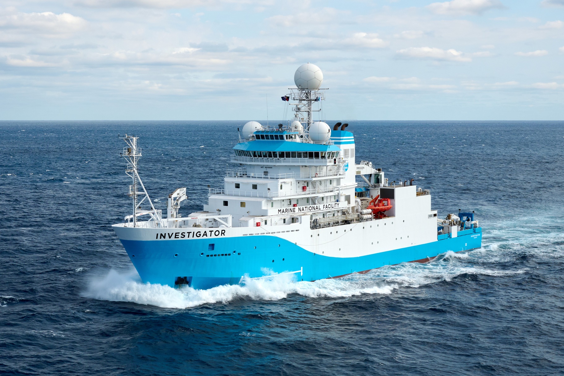 A blue and white ship named the Investigator powering through waves with a clear blue sky in the background.