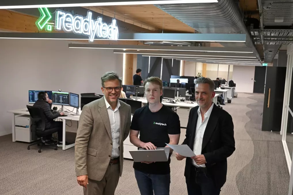 Three people in office with 'readytech' sign; two in suits, one in t-shirt holding laptop.