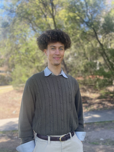 Alex Tomkinson standing outdoors on a paved path with trees and greenery in the background, wearing a dark cable-knit sweater over a collared shirt and light-colored pants.