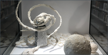 The image shows a small display case with several white, textured sculptures made from soap. There&rsquo;s a metal cheese grater standing upright with a spiralling, ribbon like soap sculpture swirling out of its top. On the left is a coral shaped piece, and on the right is a rough white sphere. The surface is scattered with small round white soap discs. In front, there&rsquo;s a black &ldquo;Please Do Not Disturb&rdquo; door hanger. The background is a library shelf seen through glass.