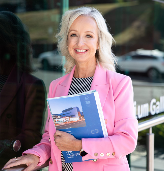 Vaness Cahoon wearing a pink blazer stands outdoors by a glass building, holding a blue booklet featuring a building photo and the text “Don McTaggart Society,” with a blurred street and railing in the background.