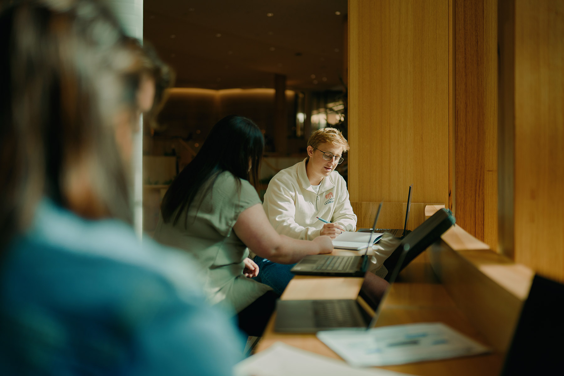 Students studying together in the new Forestry building, using laptops at a window-side workspace with warm ambient lighting and timber interiors.