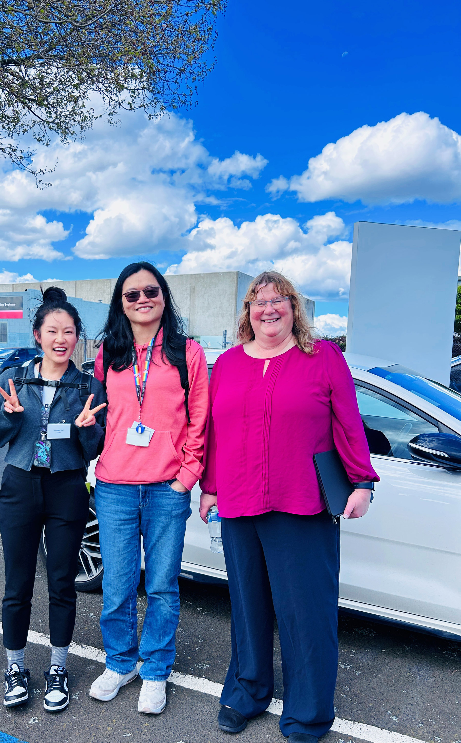 Luoyan and her friends standing beside a white car in a car park, wearing visitor lanyards.