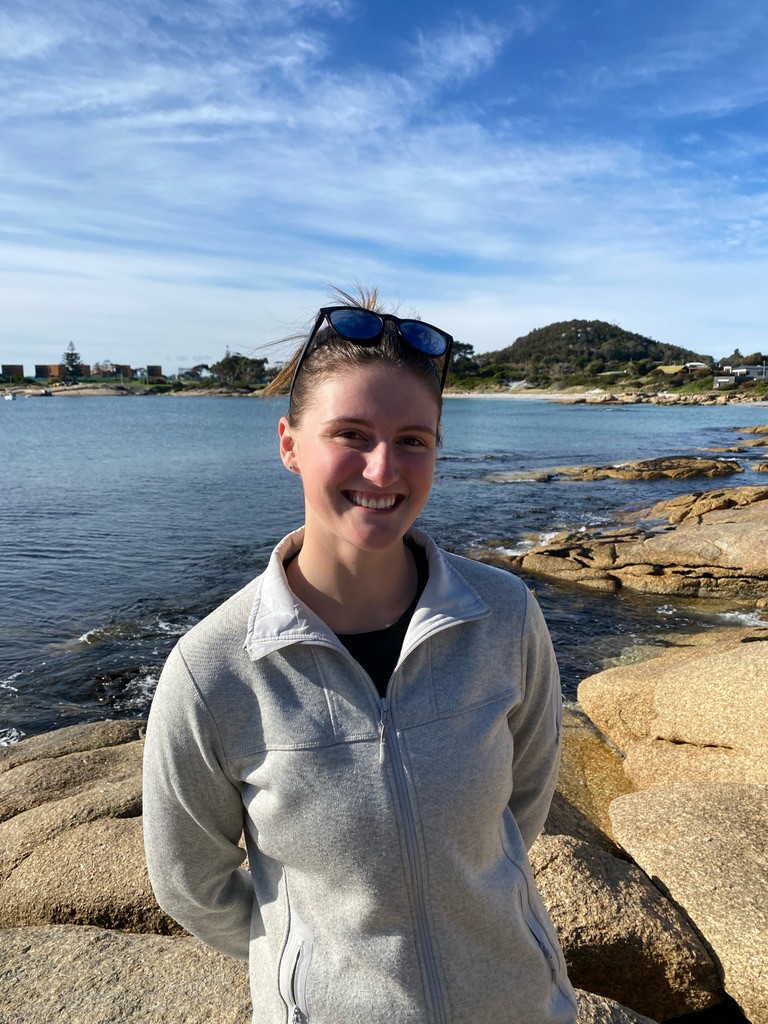 A young woman in a grey zip-up jumper, sunglasses on top of her head, stands smiling on the rocky shore of a Tasmanian beach