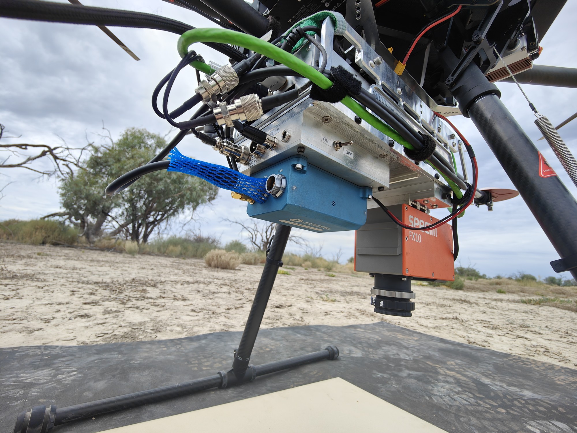 Close-up of drone underside with wiring and components, resting outdoors near trees and bushes.