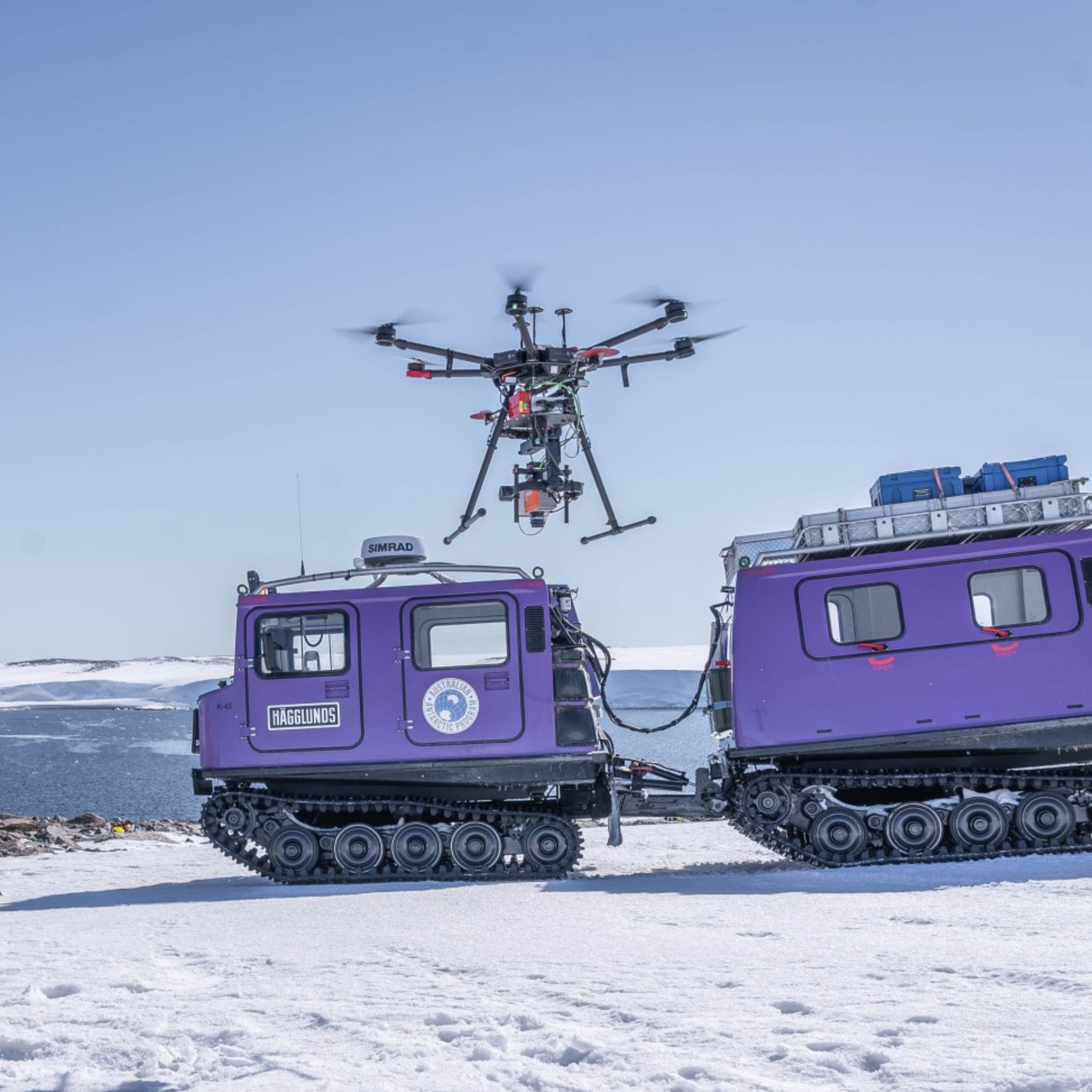 Purple tracked vehicle towing trailer in snow with drone flying above, carrying equipment.