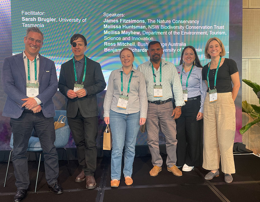 Six panelists with conference badges stand in front of a screen listing their names and affiliations at the ALCA conference..