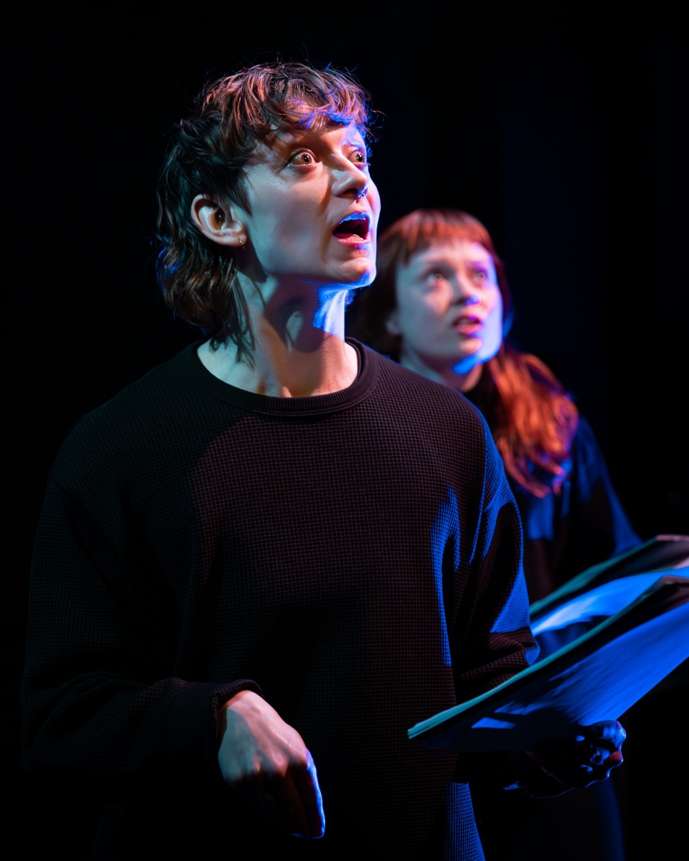 Close up of Two women on stage, one at the forefront with short dark hair and a black top holding a script