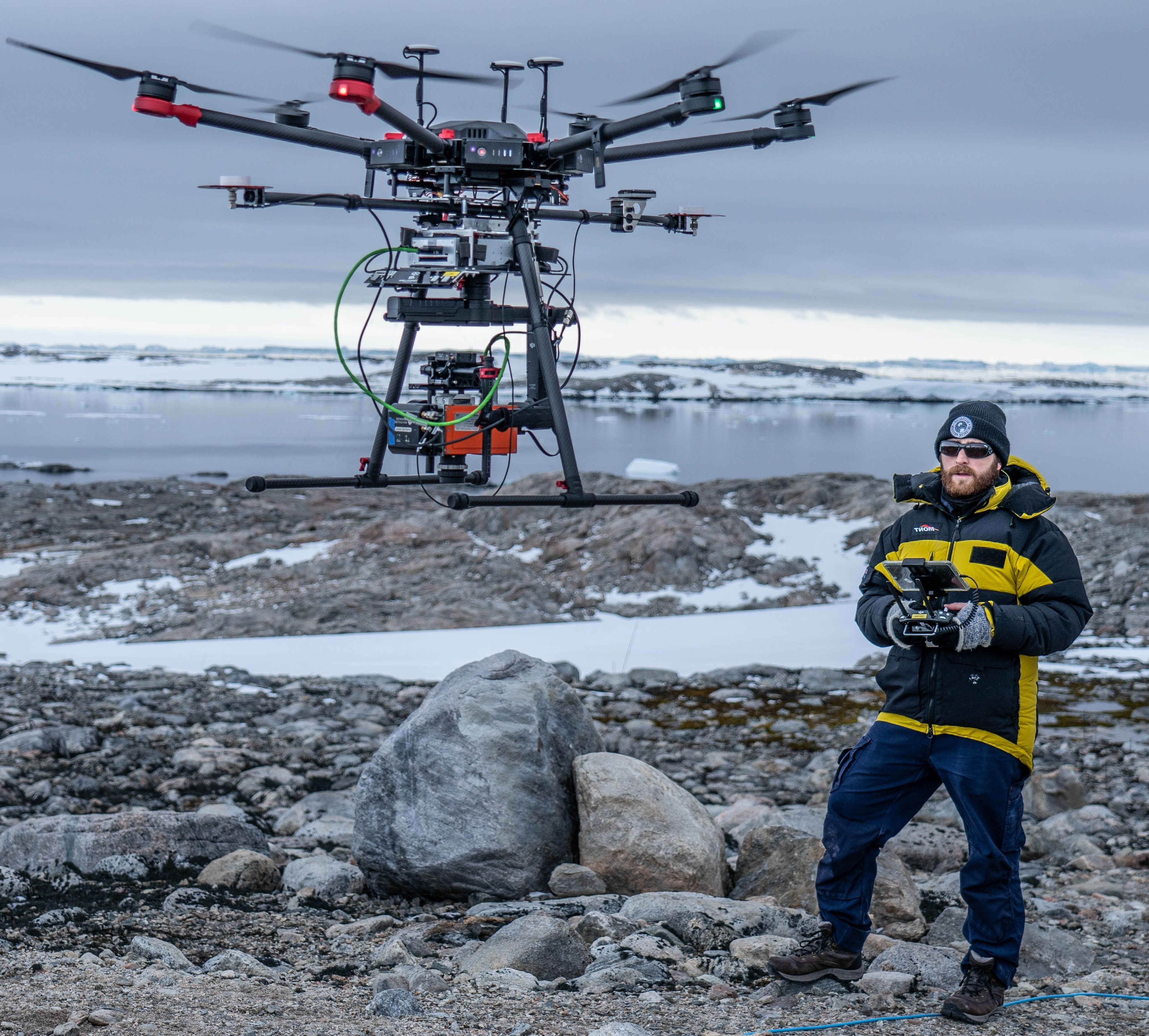 Ryan Haynes in a yellow and black hooded winter jacket operating a large multi-rotor drone in a rocky, snowy area near a body of water, with an overcast sky.