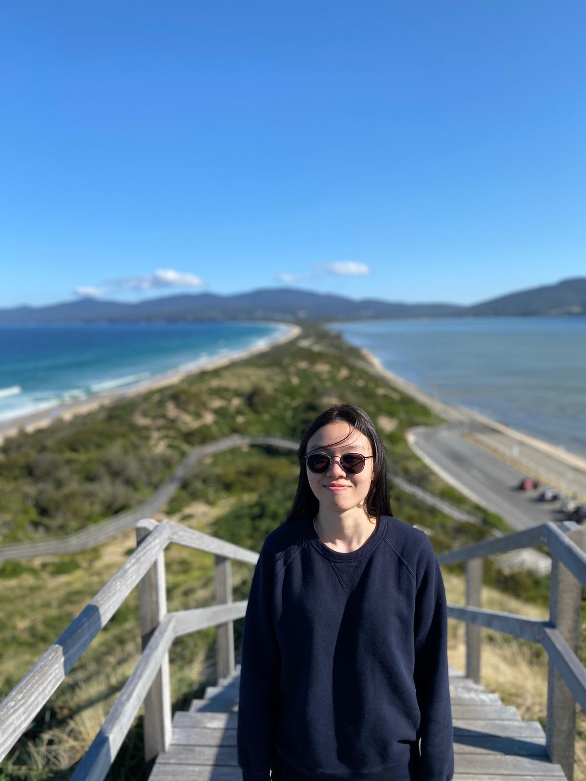 A student in a dark jumper and sunglasses stands on top of a tall wooden structure. In the background are two bodies of water divided by a thin strip of land.