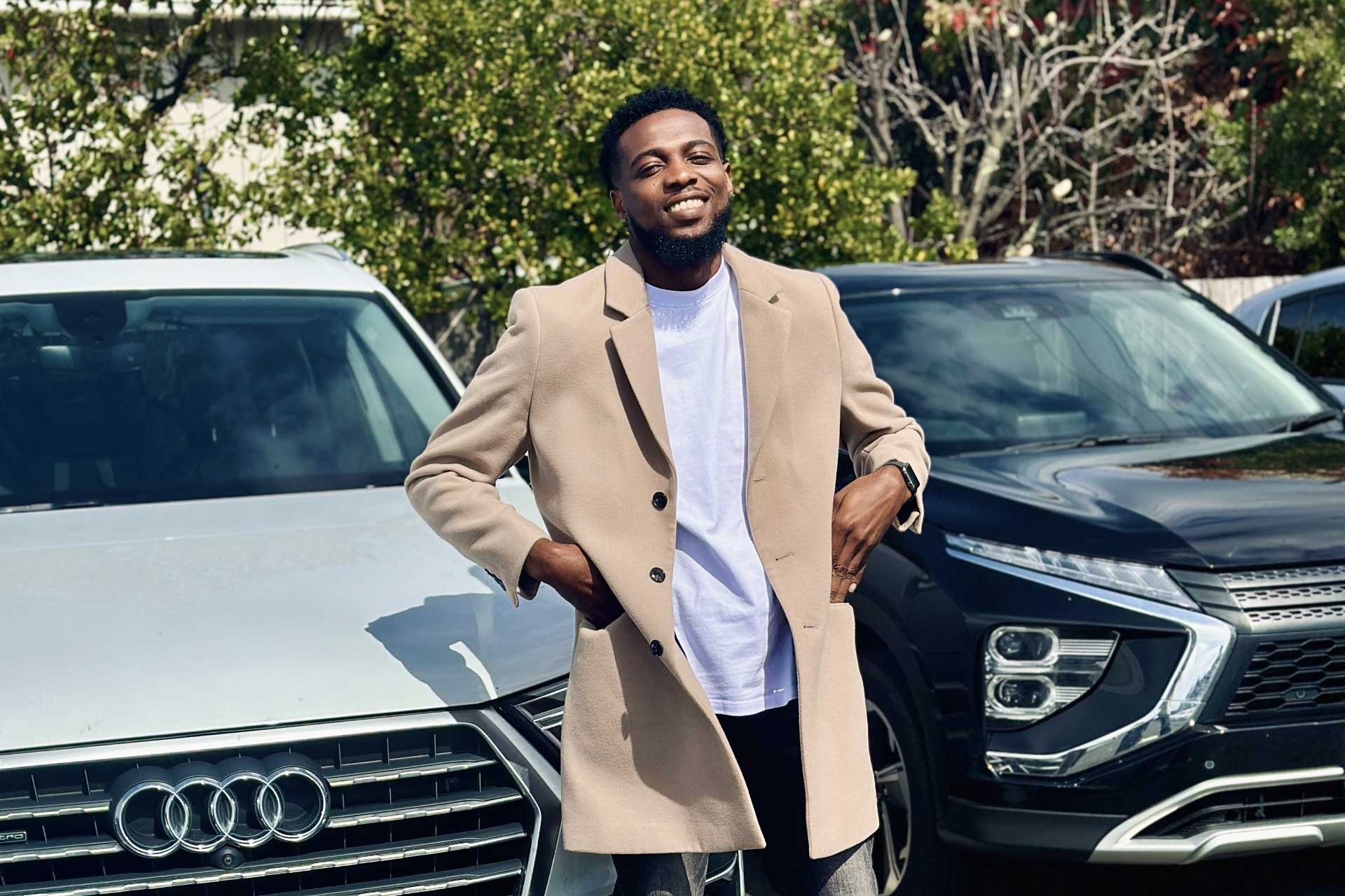 Person in beige coat standing between an Audi and a Mitsubishi, with trees and bushes in background.
